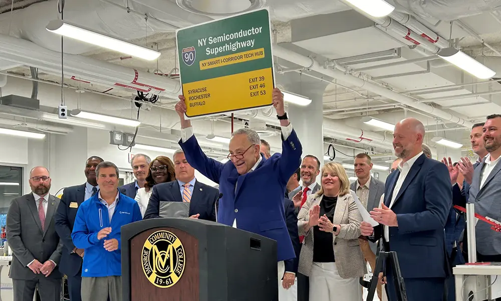 Tom Golisano smiles while holding a folder as people mingle around him.